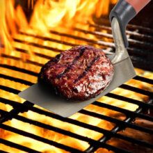 Juicy burger with appetizing grill marks on a silver spatula over a flaming charcoal barbecue grill